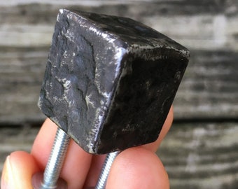 May include: A dark gray, cube-shaped metal cabinet knob with a textured, uneven surface and a rubbed hot beeswax finish. The knob features two silver-colored threaded metal bolts extending from the bottom for mounting purposes.