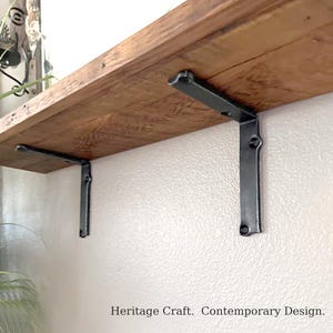 May include: A natural wood shelf mounted to a wall with two dark gray metal L-shaped brackets. The wood has a visible grain and texture. The text Heritage Craft. Contemporary Design. is printed at the bottom of the image.