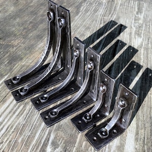 May include: A collection of six dark gray, hand-forged metal shelf brackets. Each bracket has a rectangular base with pre-drilled holes, a curved support arm, and a decorative loop. The brackets are arranged on a weathered wooden surface.