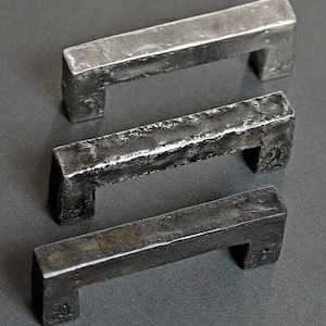 May include: Three rectangular metal drawer pulls with a dark, hammered, and textured finish. Each handle features a straight bar design with two perpendicular mounting posts, showcasing a raw, industrial appearance with visible surface variations.