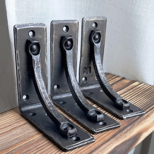 May include: Three L-shaped metal shelf brackets with a dark, hammered finish. Each bracket features a vertical back plate and a horizontal base plate connected by a curved, decorative support arm. Pre-drilled holes are visible for mounting.