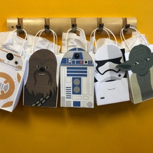 Puede incluir: Cinco bolsas de regalo de papel blanco con diferentes personajes de Star Wars impresos en ellas. Los personajes son R2-D2, Chewbacca, Stormtrooper, Yoda y BB-8. Las bolsas est&aacute;n colgadas en un estante de madera.
