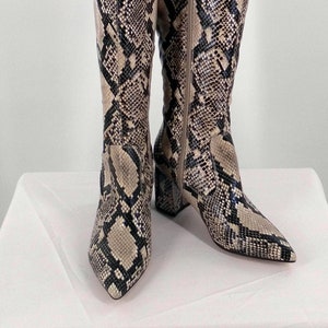 Peut inclure: Une paire de bottes mi-hautes à motif serpent, dans des tons de brun et de noir. Les bottes ont une pointe pointue et un talon bloc.