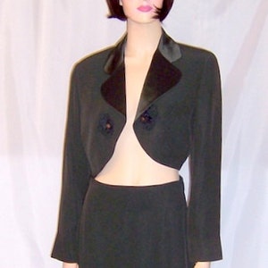 Op de afbeelding: Een zwart getailleerd jasje met een satijnen kraag en twee zwarte bloemversieringen. Het jasje wordt gecombineerd met een bijpassende zwarte rok met een split aan de voorkant.