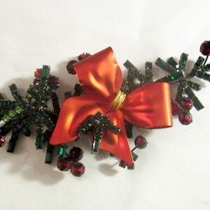 May include: A red bow with gold trim is centered on a cluster of green and black sparkly pine branches with red berry accents. This is a Christmas-themed brooch.