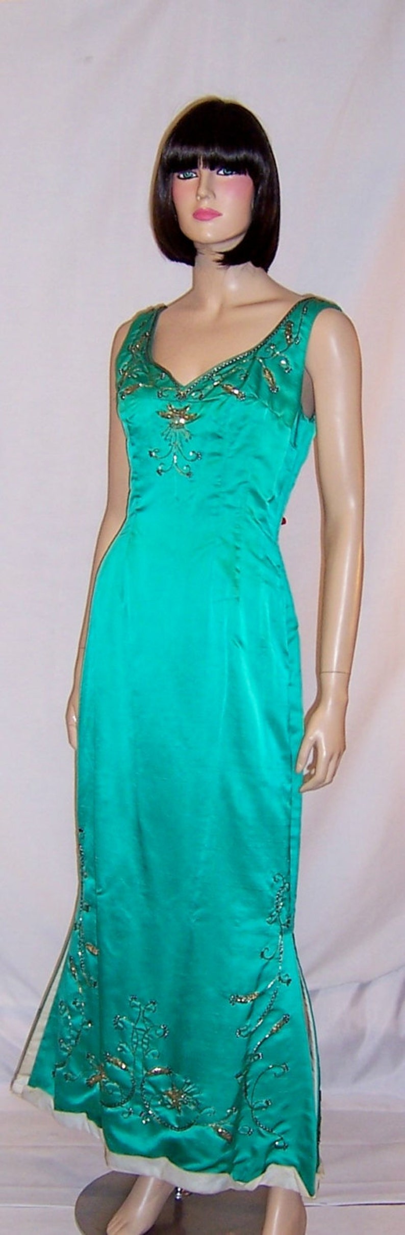 May include: A teal satin evening gown with gold and silver embellishments. The gown has a V-neckline and a fitted bodice. The skirt is full-length and has a slight train.