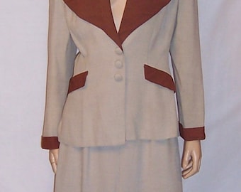 May include: A beige suit with a brown collar and brown trim on the sleeves. The suit jacket has a V-neck and four buttons down the front. The skirt is knee-length and has a slit in the back.