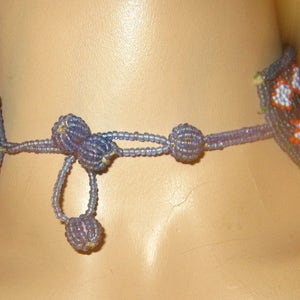 以下が含まれることがあります： 青と白のパターンでビーズでできたネックレス。ネックレスは後ろで結ばれており、端にタッセルが付いています。