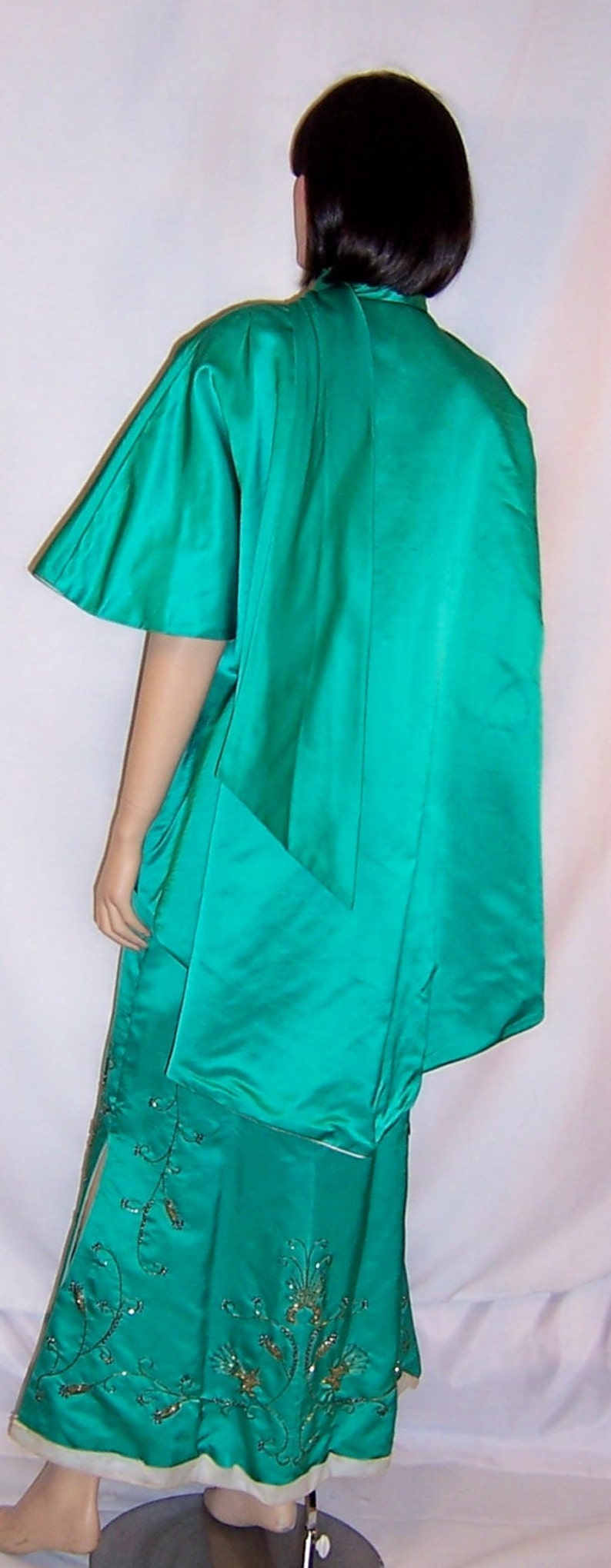May include: A teal green satin dress with a long, flowing cape. The dress has a fitted bodice and a full skirt with gold embroidery.