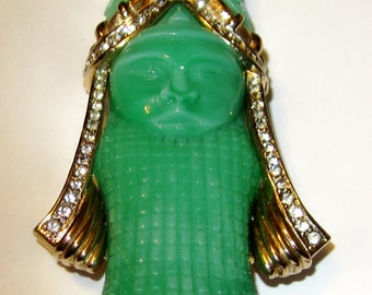 May include: A green glass brooch with a face and a gold crown. The brooch is decorated with rhinestones.
