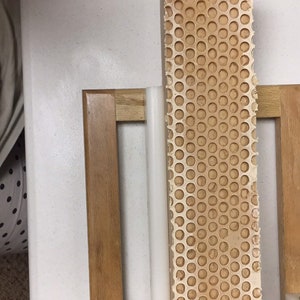 Puede incluir: Un marco de madera marr&oacute;n claro con un bloque rectangular perforado. El bloque tiene un patr&oacute;n de panal de peque&ntilde;os agujeros espaciados uniformemente. Un objeto cil&iacute;ndrico blanco est&aacute; posicionado junto al bloque.