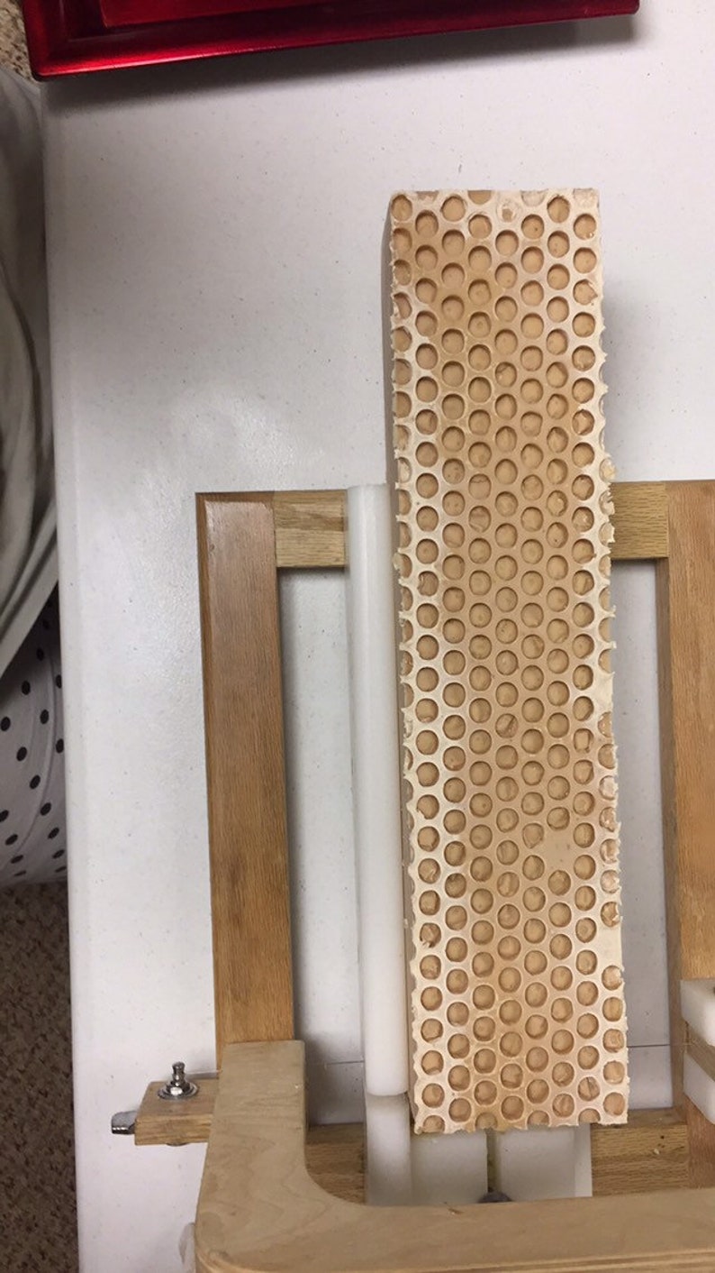 Puede incluir: Un marco de madera marr&oacute;n claro con un bloque rectangular perforado. El bloque tiene un patr&oacute;n de panal de peque&ntilde;os agujeros espaciados uniformemente. Un objeto cil&iacute;ndrico blanco est&aacute; posicionado junto al bloque.