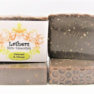 Puede incluir: Cuatro barras de jab&oacute;n apiladas, dos envueltas en pl&aacute;stico transparente. El jab&oacute;n es de color marr&oacute;n oscuro con motas visibles. Una barra tiene una etiqueta que dice "Lathers Skin Essentials Oatmeal & Honey".