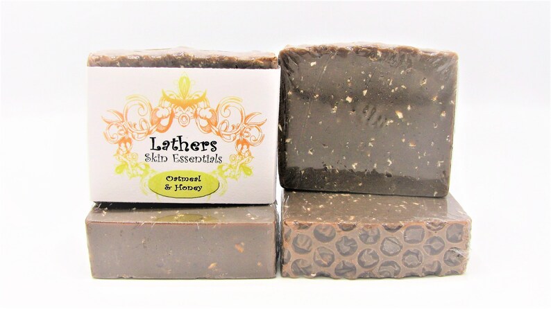 Puede incluir: Cuatro barras de jab&oacute;n apiladas, dos envueltas en pl&aacute;stico transparente. El jab&oacute;n es de color marr&oacute;n oscuro con motas visibles. Una barra tiene una etiqueta que dice "Lathers Skin Essentials Oatmeal & Honey".