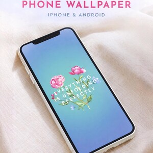 May include: A smartphone with a white frame displays a floral phone wallpaper. The wallpaper features pink peonies and the text "Everything is unfolding perfectly" on a blue gradient background. The top of the phone has a black camera.