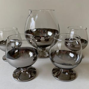 May include: A set of five clear brandy snifters with a metallic silver-colored base. The largest glass has a wide, rounded bowl and a short stem. The smaller glasses have a similar design, perfect for serving brandy or other spirits.