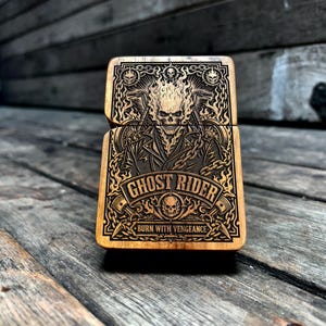 Puede incluir: Una funda rectangular de madera para encendedor con un diseño grabado a láser detallado de un personaje con calavera en llamas. El arte incluye cadenas, manillares de motocicleta y el texto GHOST RIDER y BURN WITH VENGEANCE en negrita.