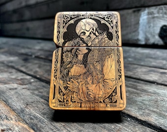 May include: A wooden lighter with an engraved design of a skeleton and a woman embracing. The design is in black and white and has a gothic aesthetic.