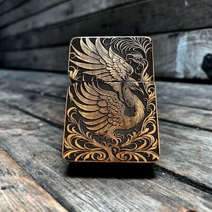 May include: A rectangular, hinged lighter with an engraved phoenix design. The lighter has a dark background with a gold phoenix and floral patterns. The metal lighter sits on a wooden surface. The design is intricate and detailed.