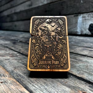 May include: A rectangular wooden lighter case featuring a detailed laser-engraved design of a Tyrannosaurus Rex, a park gate, and a vehicle. The text Jurassic Park is prominently displayed at the bottom, surrounded by intricate foliage and patterns.