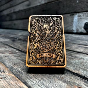 Peut inclure: Un étui à briquet rectangulaire en bois orné d'une gravure laser détaillée représentant un phénix s'élevant des flammes. Le motif comprend des volutes complexes, un soleil rayonnant et une bannière avec le mot PHOENIX en majuscules.