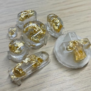 Może przedstawiać: Zestaw przezroczystych, odlewanych z żywicy guzików i elementów, każdy z zatopionym złotym listkiem. Zestaw zawiera różne kształty, takie jak zaokrąglone i prostokątne elementy oraz guzik w kształcie krzyża, wszystkie o przezroczystym wyglądzie.