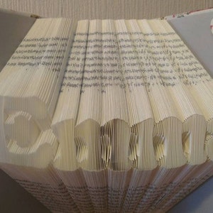 Op de afbeelding: Een handgemaakte boeksculptuur met de naam "Emma" uitgesneden uit de pagina's. Het boek is open, met de pagina's gevouwen om de letters te vormen. De kaft van het boek heeft een bloemenpatroon en een kleine hartvormige versiering.