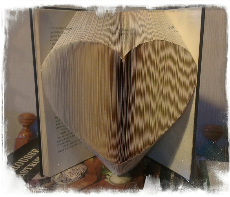 K&ouml;nnte beinhalten: Ein Buch, dessen Seiten zu einer Herzform gefaltet sind. Das Herz ist der Mittelpunkt, geformt aus den Seiten des Buches. Das offene Buch zeigt seine Seiten und erzeugt einen texturierten Effekt. Das Buch liegt auf einer Oberfl&auml;che.