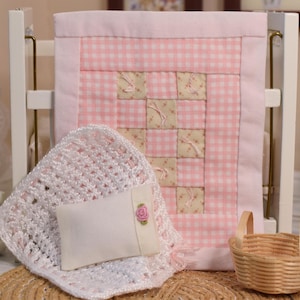 May include: A miniature bedding set featuring a pink and white gingham quilt with a central patchwork panel of floral squares, a white crocheted blanket, and a small rectangular white pillow accented with a single pink fabric rose embellishment.