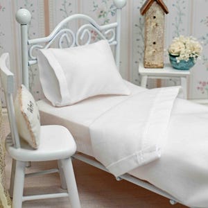 May include: White doll bed with a white metal frame, white bedding, and a white pillow. The bed is in a dollhouse room with white walls and floral wallpaper.
