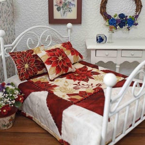 May include: Miniature white metal bed with a floral quilt and matching pillows. The quilt features a red poinsettia design on a cream background, with red and white accents. A small white nightstand and floral arrangement complete the scene.