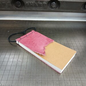 Puede incluir: Un pequeño cuaderno encuadernado en cuero rosa y marrón con un borde deshilachado. El cuaderno está atado con un cordón negro.