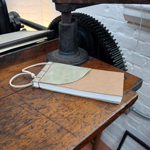 Puede incluir: Un diario hecho a mano con una cubierta de cuero verde claro y una cubierta de papel marrón, encuadernado con hilo rojo y una cuerda color crema. El diario descansa sobre una mesa de madera desgastada, cerca de una imprenta.