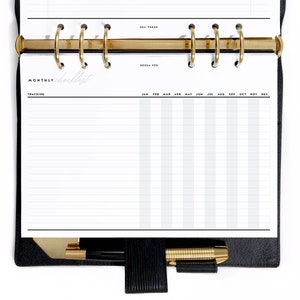 Pode incluir: Uma agenda encadernada em couro preto com argolas douradas e uma página branca com o texto "MONTHLY checklist" e uma grade para acompanhar as tarefas.