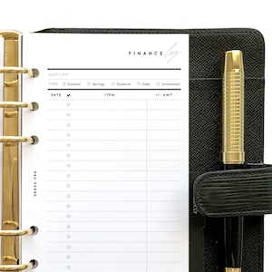 May include: A white page with a black and white grid for tracking finances. The page is labeled "FINANCE" and has columns for date, item, type, account/category, and amount. The page is in a black leather binder with a gold pen.