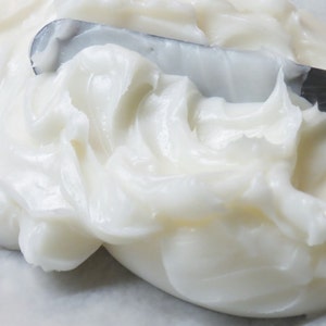 May include: A close-up of a white, creamy substance being spread with a metal spatula. The substance is smooth and appears to be a type of lotion or cream.