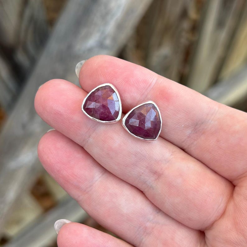 May include: A pair of ruby-colored gemstone stud earrings. Each earring features a triangular, faceted ruby stone set in a silver-colored bezel. The earrings are displayed on a person's hand.