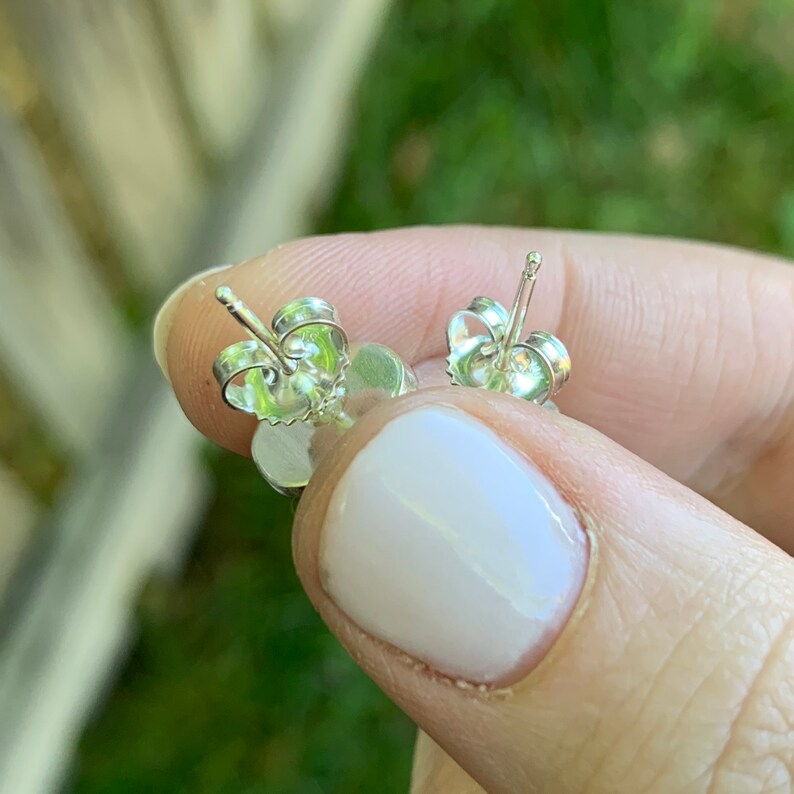 May include: Close-up of two silver stud earrings with a flower design. The earrings have a post and butterfly backing. The earrings are held up against a blurred green background.