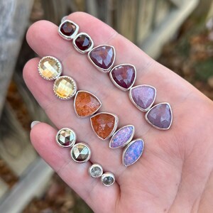 May include: A collection of gemstone stud earrings in various shapes and colors. The earrings include round, square, and triangular stones in shades of red, yellow, orange, purple, and iridescent blue. Each earring is set in a silver bezel.