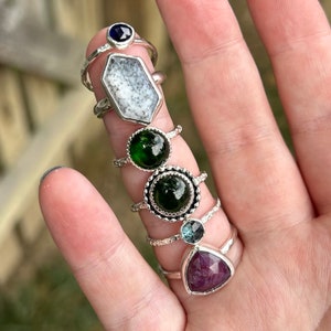 May include: A stack of silver rings with various gemstones, including a white opaque stone, a dark blue stone, two green stones, a light blue stone, and a pink stone.