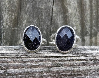 Puede incluir: Dos anillos de plata con piedras ovaladas de color azul oscuro. Las piedras tienen una textura brillante y reluciente.