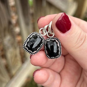 Puede incluir: Un par de pendientes de plata con una piedra preciosa negra rectangular. Las piedras preciosas están engastadas en un marco plateado con un borde punteado. Los pendientes cuelgan de un gancho plateado.