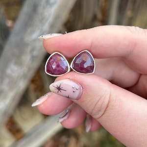 May include: A pair of triangular ruby earrings with a silver setting. The earrings feature faceted, deep red gemstones. The earrings are held up by a hand, with a blurred background.