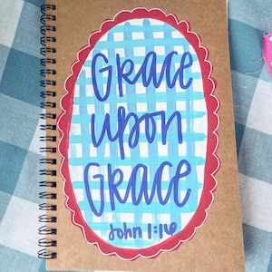 May include: A spiral-bound notebook with a brown kraft paper cover featuring a hand-painted oval design. The oval has a red scalloped border, a light blue and white checkered background, and the dark blue text "Grace upon Grace John 1:16".