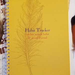 Może przedstawiać: Żółty dziennik śledzenia nawyków z ilustracją brązowego pióra i tekstem "Habit Tracker twelve month habit & mood journal".