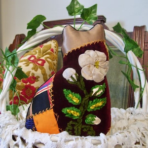 May include: A stuffed cat-shaped ornament featuring a deep burgundy velvet front panel adorned with an embroidered white flower and green leaves. The side is constructed from patchwork fabric in orange, blue, and black, joined by visible yellow stitching.