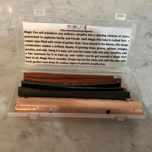 K&ouml;nnte beinhalten: Eine transparente Kunststoffbox mit einem Kupferrohr und mehreren Materialstreifen. Die Box hat ein Etikett mit dem Text "MAGIC FIRE" und Produktinformationen. Das Produkt soll farbenfrohe Flammen in einem Lagerfeuer erzeugen.