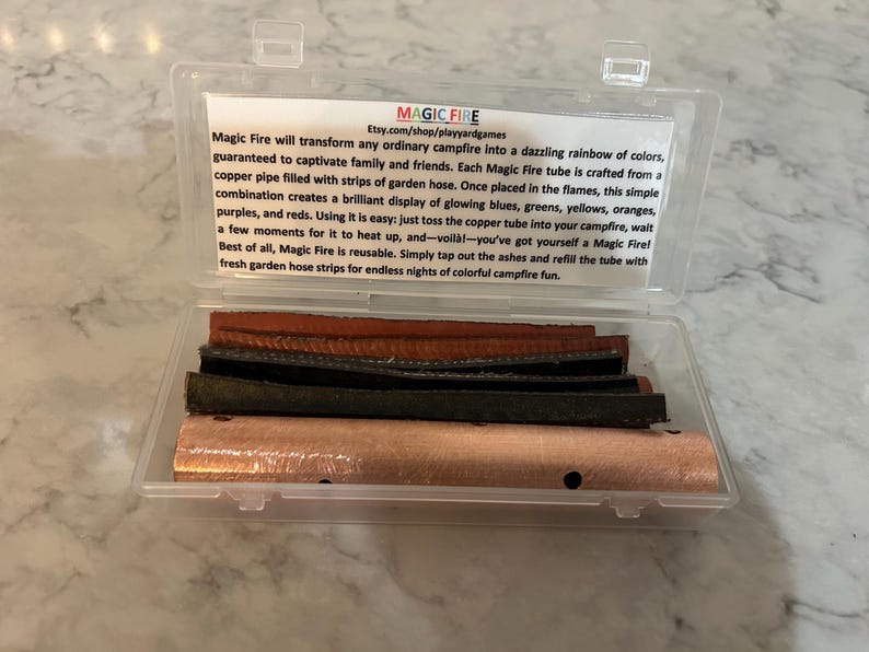 K&ouml;nnte beinhalten: Eine transparente Kunststoffbox mit einem Kupferrohr und mehreren Materialstreifen. Die Box hat ein Etikett mit dem Text "MAGIC FIRE" und Produktinformationen. Das Produkt soll farbenfrohe Flammen in einem Lagerfeuer erzeugen.