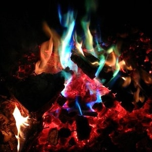K&ouml;nnte beinhalten: Ein lebendiges Lagerfeuer mit Flammen in Blau-, Gr&uuml;n-, Orange- und Rott&ouml;nen. Das brennende Holz und die Glut leuchten vor einem dunklen Hintergrund und erzeugen eine farbenfrohe und dynamische Szene.