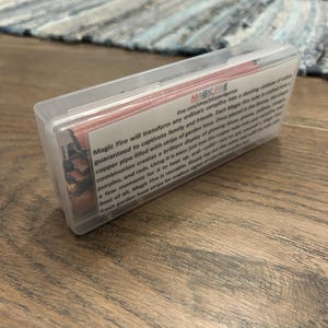 K&ouml;nnte beinhalten: Eine transparente Kunststoffbox mit Streifen aus Gartenschlauch f&uuml;r Lagerfeuer. Auf der Box steht "MAGIC FIRE" und eine Produktbeschreibung. Die Streifen erzeugen ein farbenfrohes Flammenspiel.